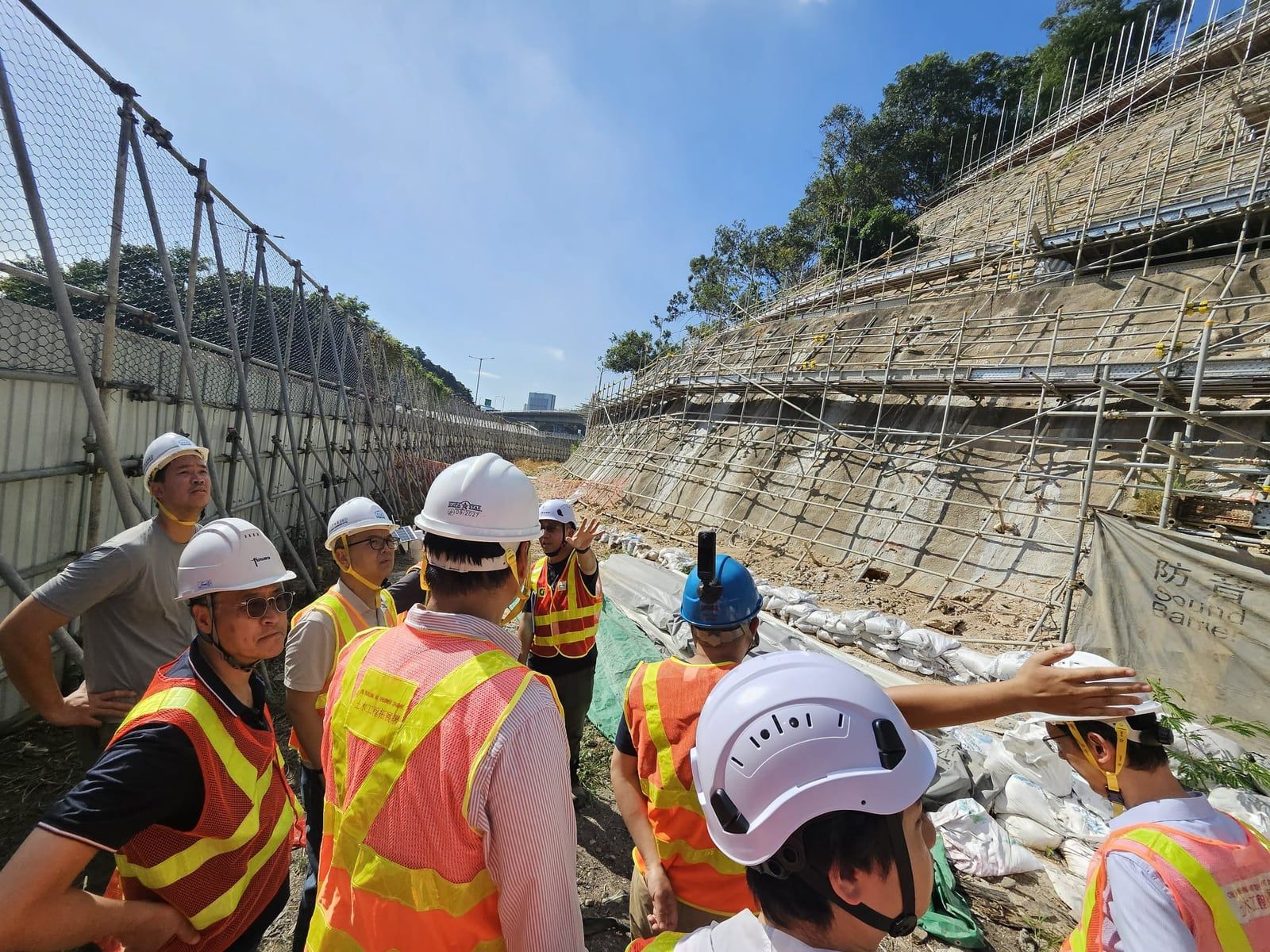 Engineers and government staff in hard hats and high-visibility vests inspect a scaffolded hillside on a Hong Kong works site